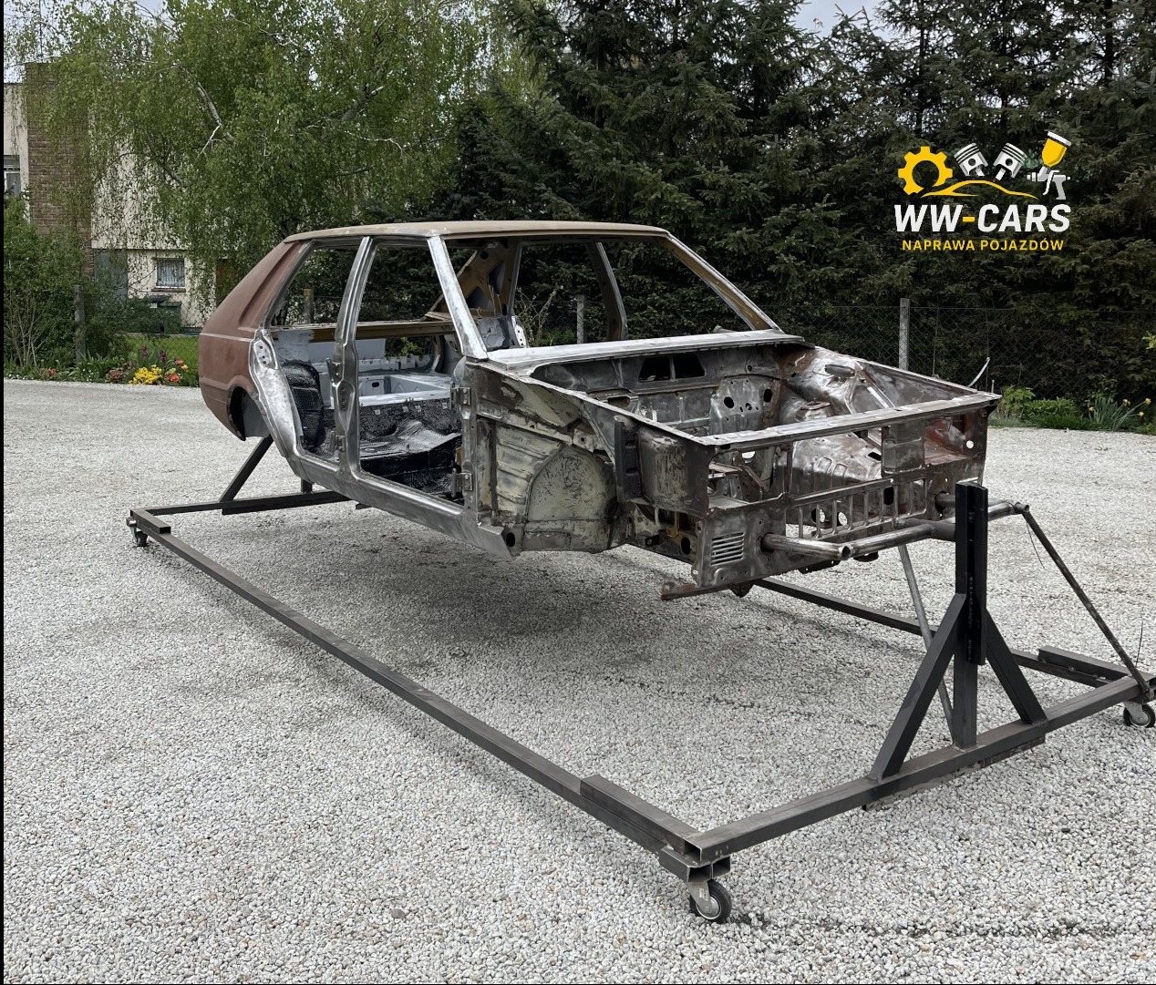 Karoseria samochodu na metalowym stelażu z kółkami, widok z boku, na zewnątrz, w tle zieleń. Częściowo zardzewiała, przygotowana do dalszych prac.