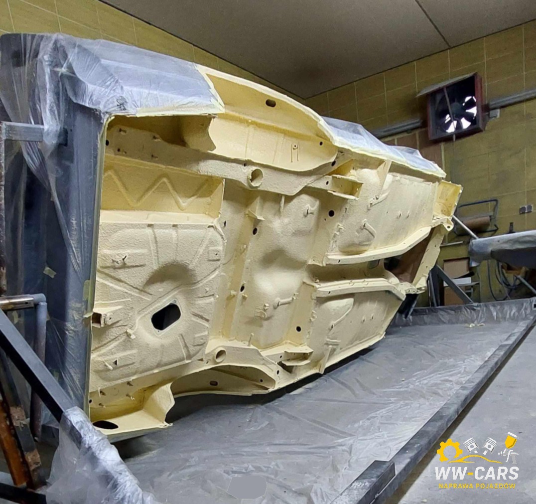 Podwozie samochodu po nałożeniu warstwy ochronnej, widoczne detale konstrukcyjne i otwory. Częściowo zakryte folią ochronną, w tle wentylator.