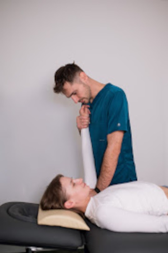 Pacjentka leży na kozetce, a specjalista w turkusowej koszulce wykonuje mobilizację stawu barkowego. Jasne tło, stonowane oświetlenie.