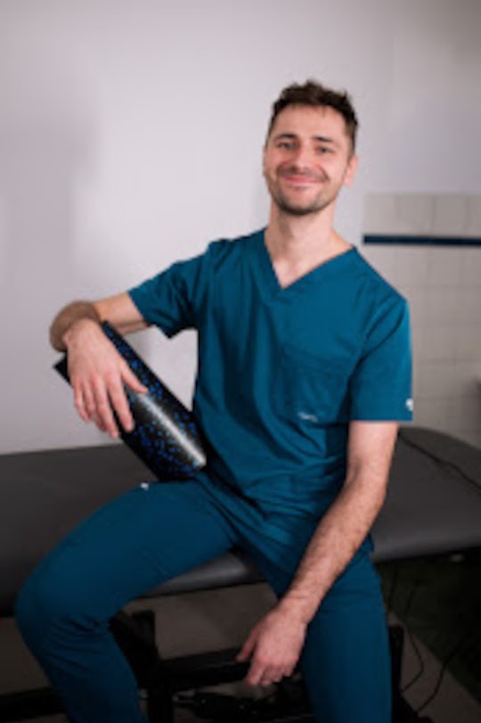 Uśmiechnięty mężczyzna w turkusowym uniformie siedzi na leżance, trzymając wałek do masażu. W tle biała ściana i fragment kafelków.