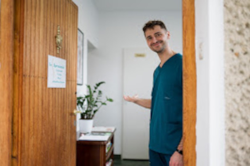 Uśmiechnięty mężczyzna w turkusowym uniformie zaprasza do gabinetu z drewnianymi drzwiami, na których wisi informacja. W tle widoczny stolik i roślina doniczkowa.