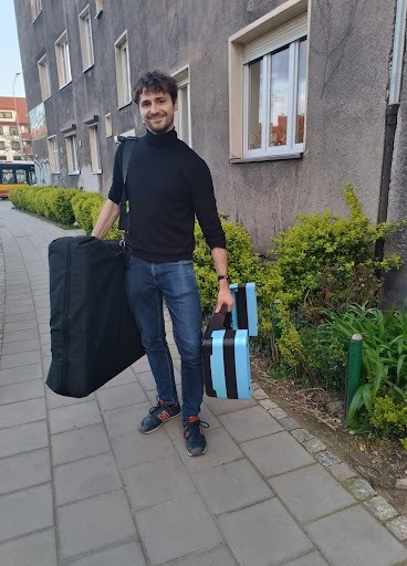 Uśmiechnięty mężczyzna w czarnym golfie i jeansach, niosący torbę i niebiesko-czarne pudełka na chodniku przed budynkiem. Gotowy do pracy.