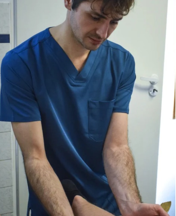 Mężczyzna w niebieskim uniformie pochyla się nad pacjentem, prawdopodobnie podczas sesji terapeutycznej. Widoczne dłonie i fragment nogi pacjenta.