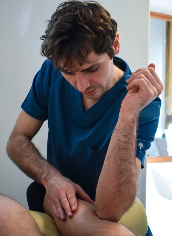 Mężczyzna w niebieskim uniformie wykonuje masaż kolana pacjenta. Widoczny fragment kozetki i dłoń terapeuty. Delikatne oświetlenie podkreśla detale.