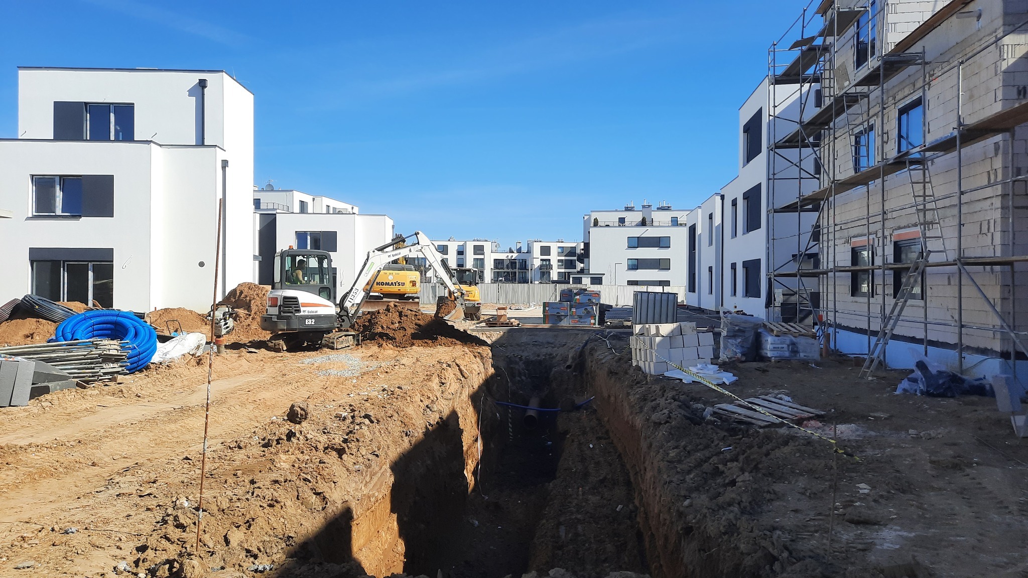 Budowa osiedla: Wykop pod fundamenty z widocznymi maszynami budowlanymi, rurami i blokami. W tle nowoczesne, białe budynki w trakcie wznoszenia.