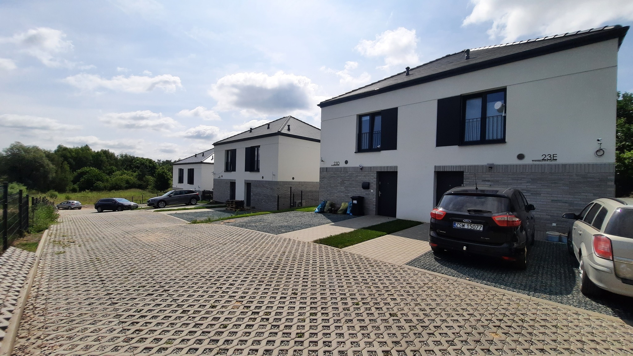 Nowoczesne, szeregowe domy z czarnymi dachami i oknami, z kostką ażurową na podjazdach i zaparkowanymi samochodami. Widok z perspektywy ulicy w słoneczny dzień.