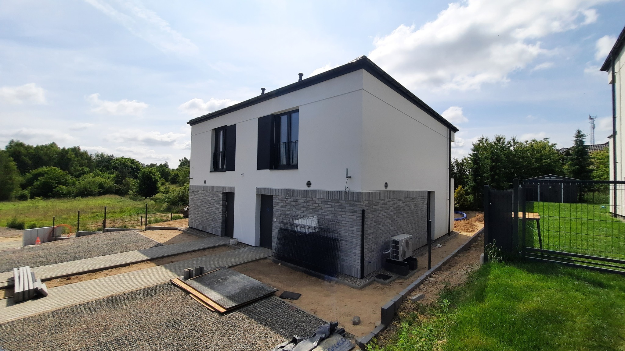 Nowoczesny, dwukondygnacyjny dom bliźniak z białymi ścianami i szarym wykończeniem z cegły, widoczny z zewnątrz w słoneczny dzień. Teren wokół domu w trakcie zagospodarowania.