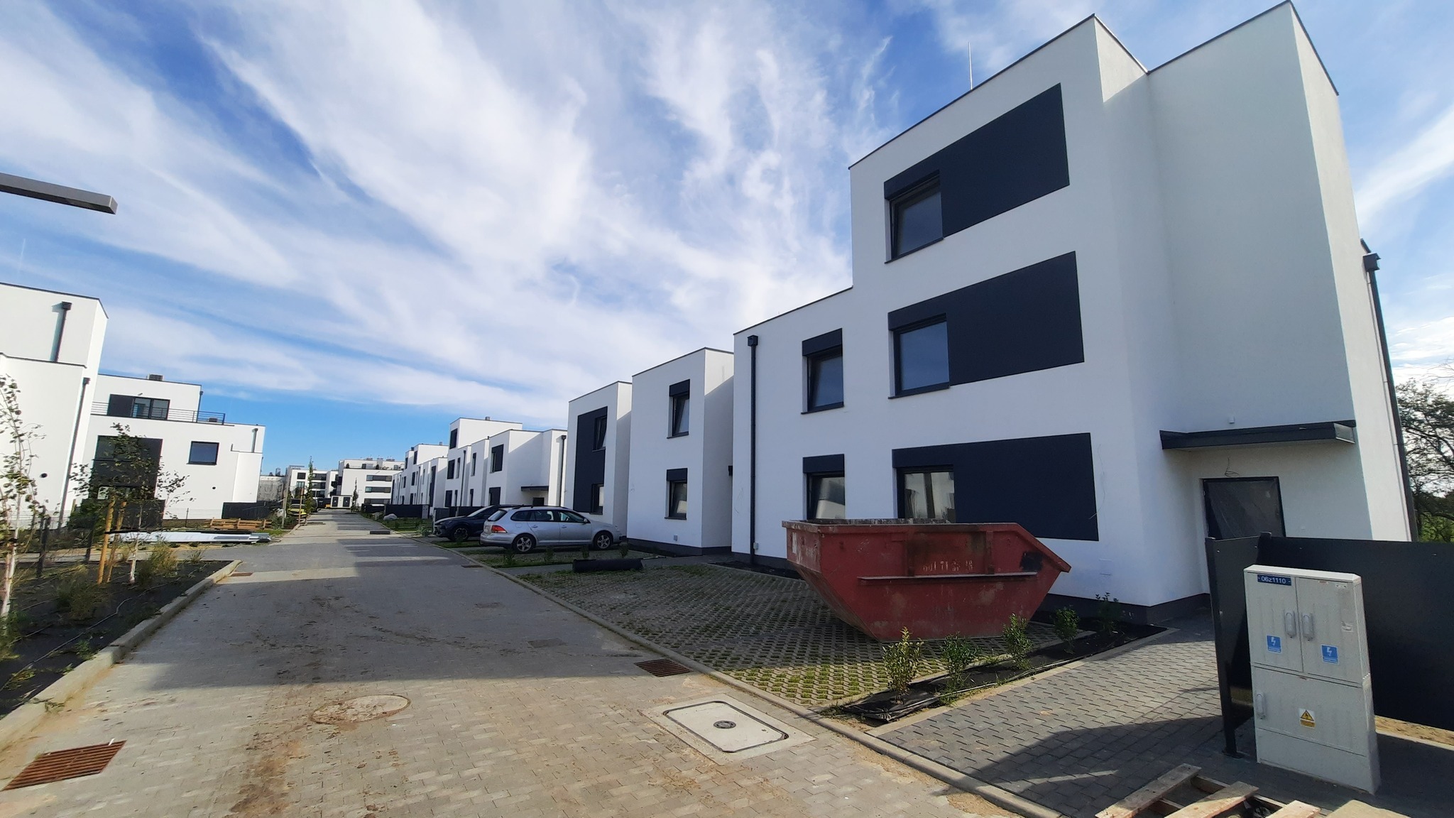 Nowoczesne, białe domy szeregowe z ciemnymi oknami i elementami elewacji, ustawione wzdłuż brukowanej drogi osiedlowej pod błękitnym niebem z chmurami.