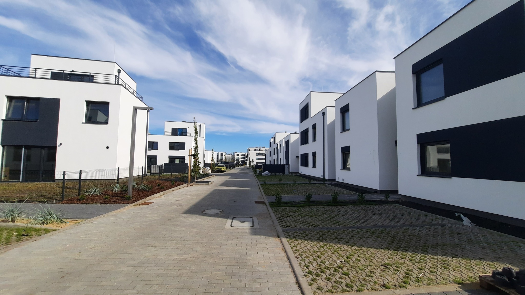 Nowoczesne osiedle domów jednorodzinnych w minimalistycznej stylistyce, z białymi elewacjami i ciemnymi akcentami, widok z perspektywy ulicy w słoneczny dzień.