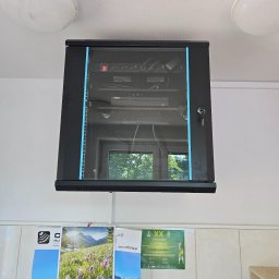 NETCAM Marcin Radochoński - Zamknięta szafa rack z urządzeniami sieciowymi, zamontowana na ścianie z kafelkami i kalendarzem. Widoczny zamek z kluczem i urządzenia Dahua wewnątrz.