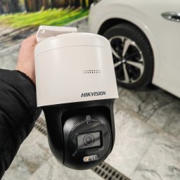 NETCAM Marcin Radochoński - Kamera obrotowa Hikvision w dłoni, na tle białego samochodu i dekoracyjnej ściany z motywem leśnej drogi. Widoczny obiektyw i logo producenta.