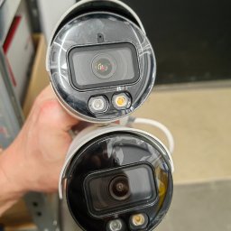 NETCAM Marcin Radochoński - Dwie kamery monitoringu w dłoni, zbliżenie na obiektywy i diody IR. Widoczne białe obudowy i czarne ramki obiektywów. Tło stanowią półki magazynowe.