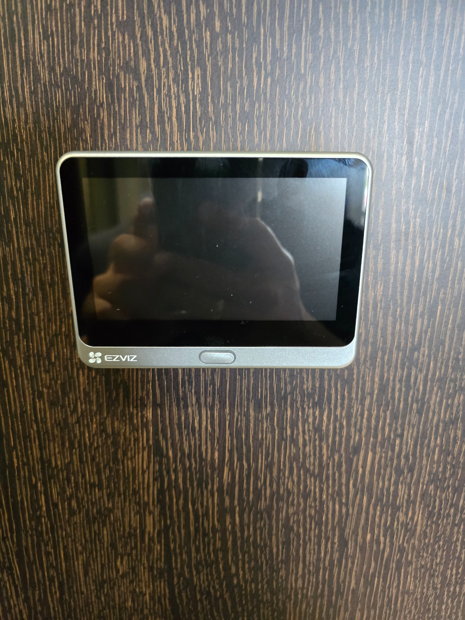 Zamontowany wideodomofon EZVIZ z ekranem na ciemnobrązowych drzwiach. Widoczny srebrny panel z logo i przyciskiem. Ekran odbija światło.