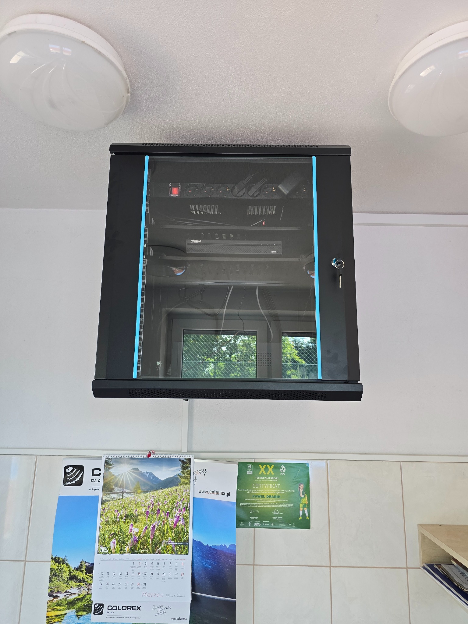 Zamknięta szafa rack z urządzeniami sieciowymi, zamontowana na ścianie z kafelkami i kalendarzem. Widoczny zamek z kluczem i urządzenia Dahua wewnątrz.