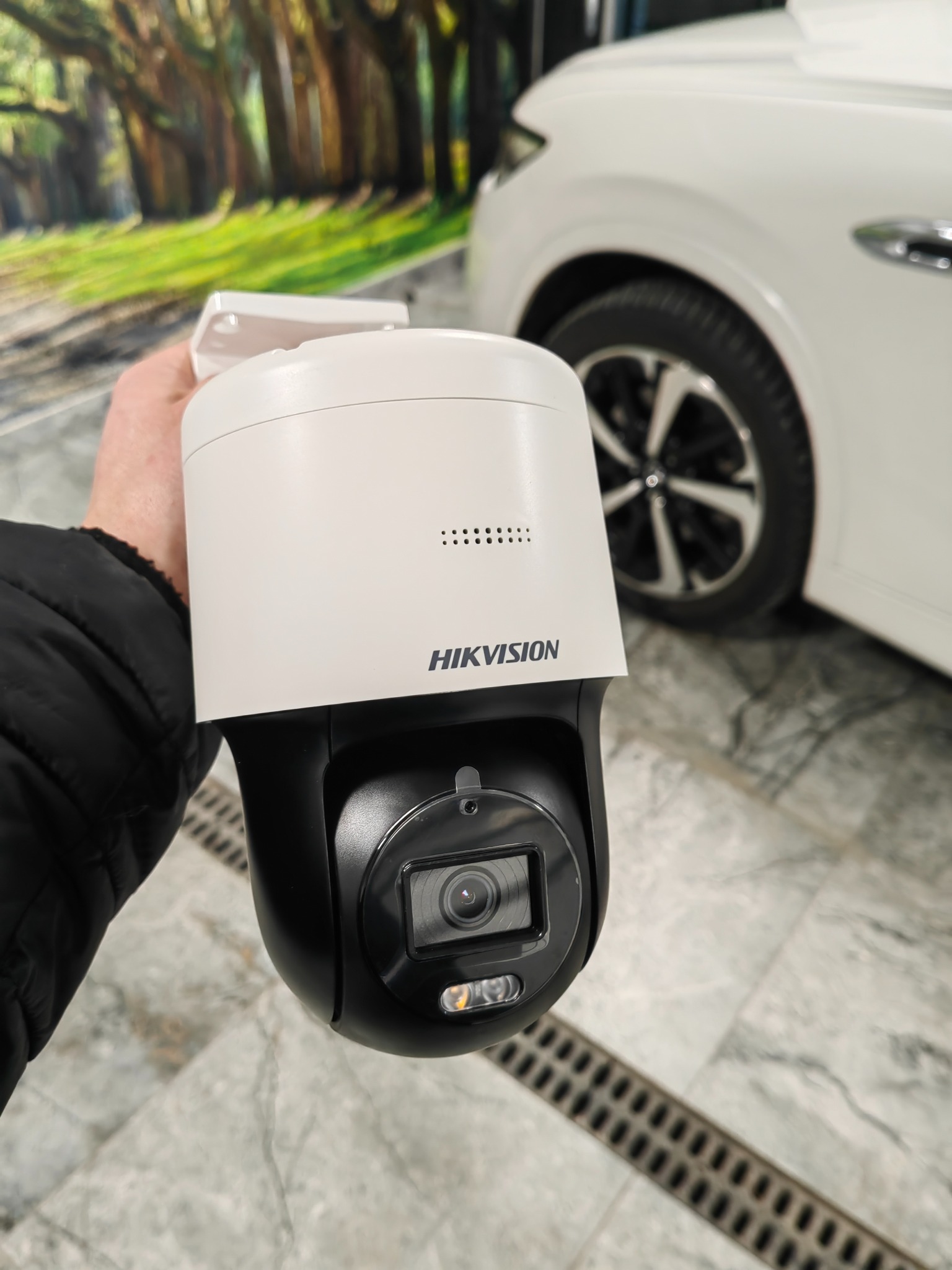 Kamera obrotowa Hikvision w dłoni, na tle białego samochodu i dekoracyjnej ściany z motywem leśnej drogi. Widoczny obiektyw i logo producenta.