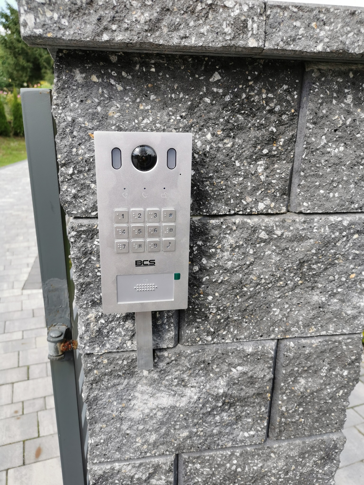 Szary panel domofonu BCS z klawiaturą numeryczną i kamerą, zamontowany na szarym, kamiennym słupku ogrodzenia. Widoczny fragment furtki.