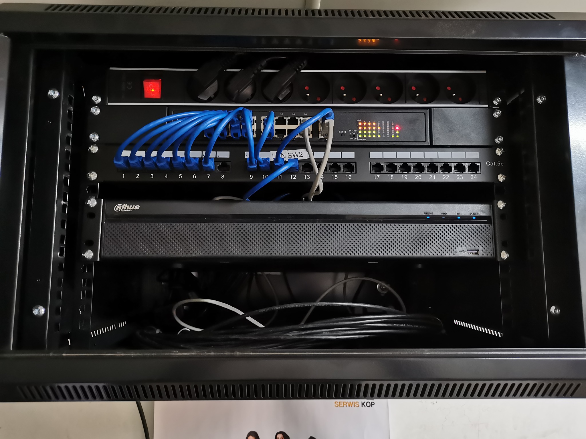 Zamknięta szafa rack z zamontowanym switchem, patch panelem i listwą zasilającą. Widoczne niebieskie kable sieciowe i czarne przewody zasilające.