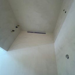 TP Dekor - Wnętrze po gładziowaniu ścian i sufitu, widoczne otwory instalacyjne i odpływ liniowy. Ściany w jasnym odcieniu, gotowe do dalszych prac wykończeniowych.