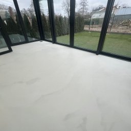 TP Dekor - Jasna, gładka posadzka w oranżerii z czarnymi ramami okiennymi. Widok na zielony trawnik i drzewa za oknem. Minimalistyczny design, neutralna kolorystyka.