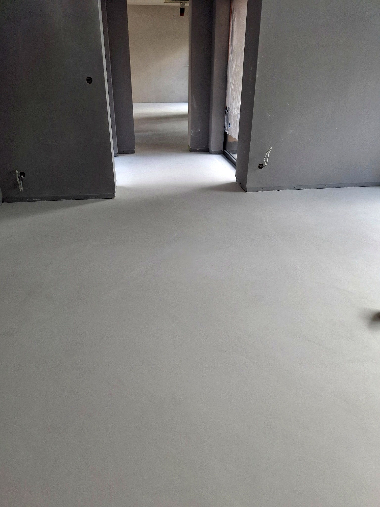 Jasnoszara, gładka posadzka w surowym wnętrzu z szarymi ścianami i otworami drzwiowymi. Minimalistyczny design, widoczne kable elektryczne.