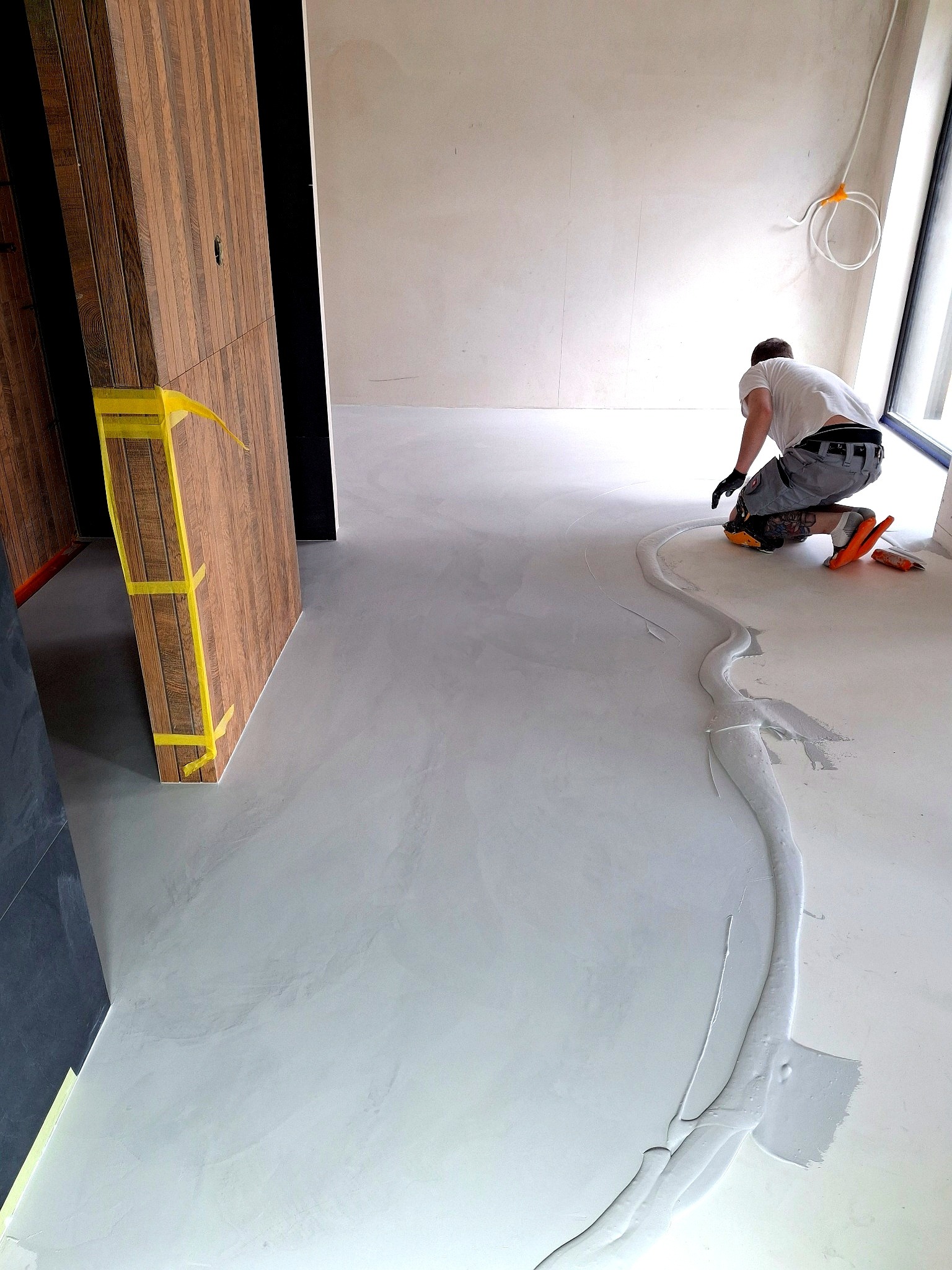 Aplikacja mikrocementu na podłodze przez wykonawcę. Widoczny fragment ściany z drewnianymi panelami zabezpieczonymi taśmą. Jasne wnętrze w trakcie wykańczania.