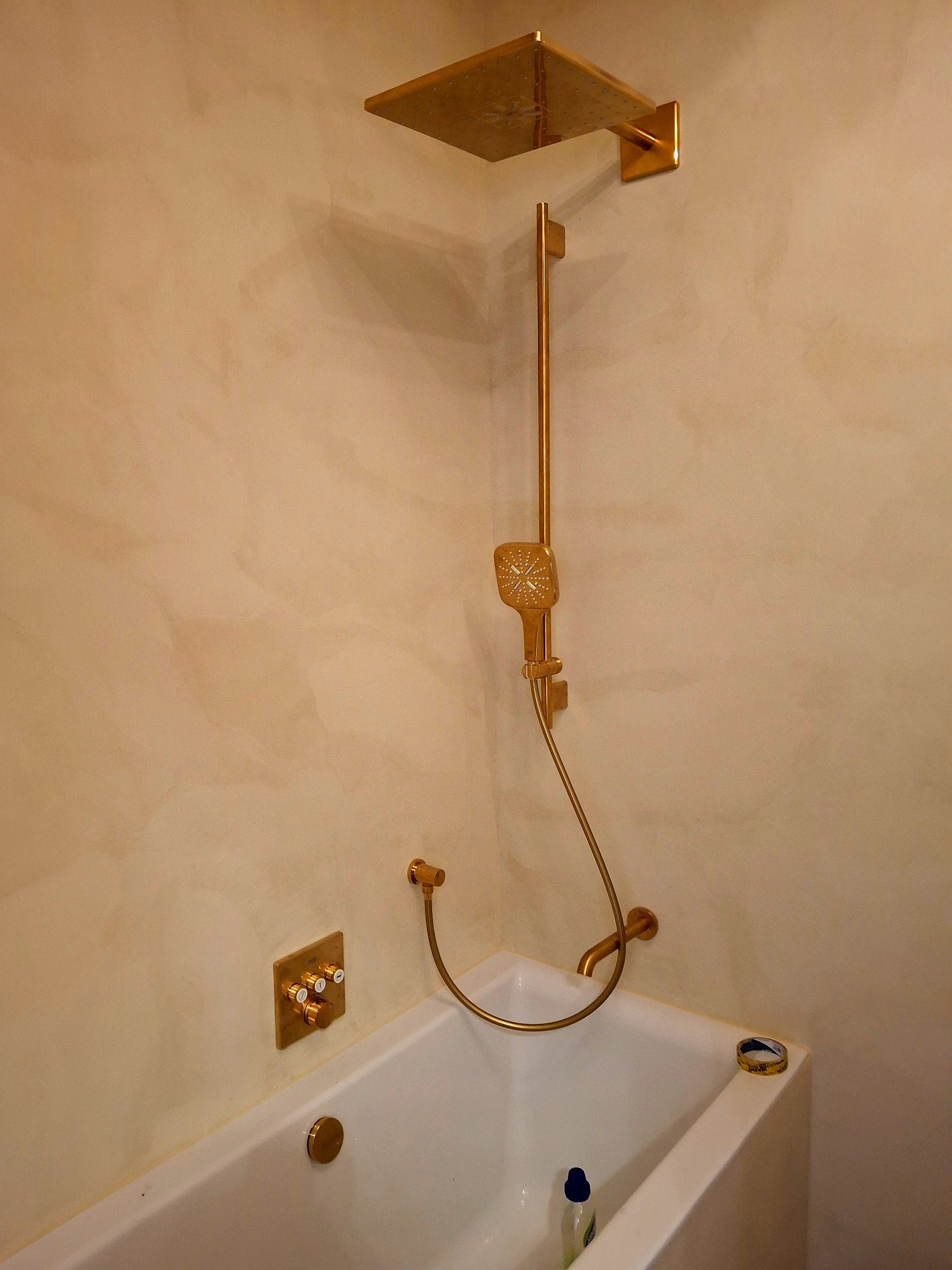 Wykończona łazienka z wanną i złotą armaturą, ściany w odcieniach beżu z delikatną fakturą. Widoczny zestaw prysznicowy i bateria wannowa.