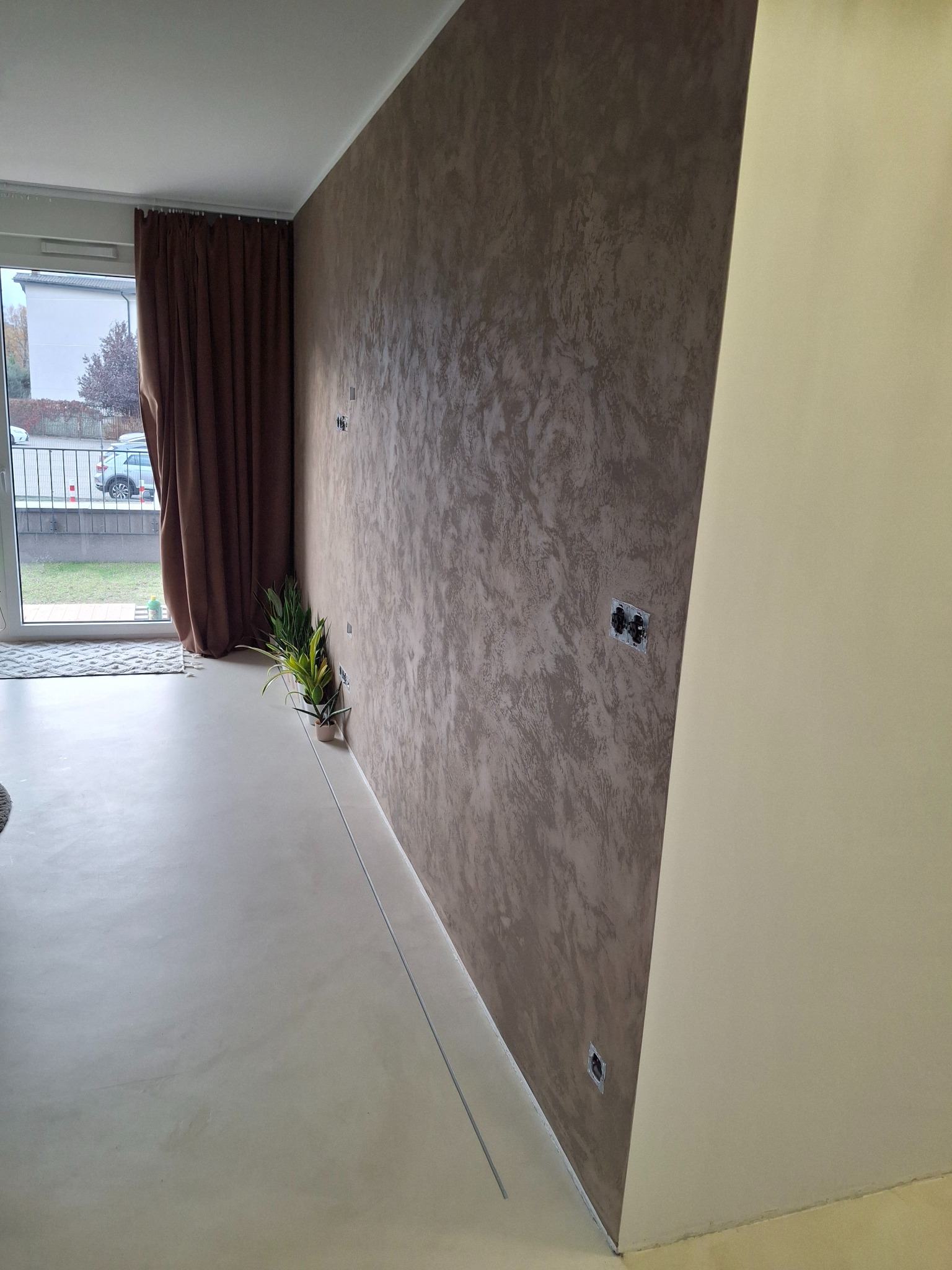 Pokój po remoncie: ściana z efektem dekoracyjnym w odcieniach brązu, jasna podłoga, okno z brązowymi zasłonami i rośliny doniczkowe. Widoczne gniazdka elektryczne.