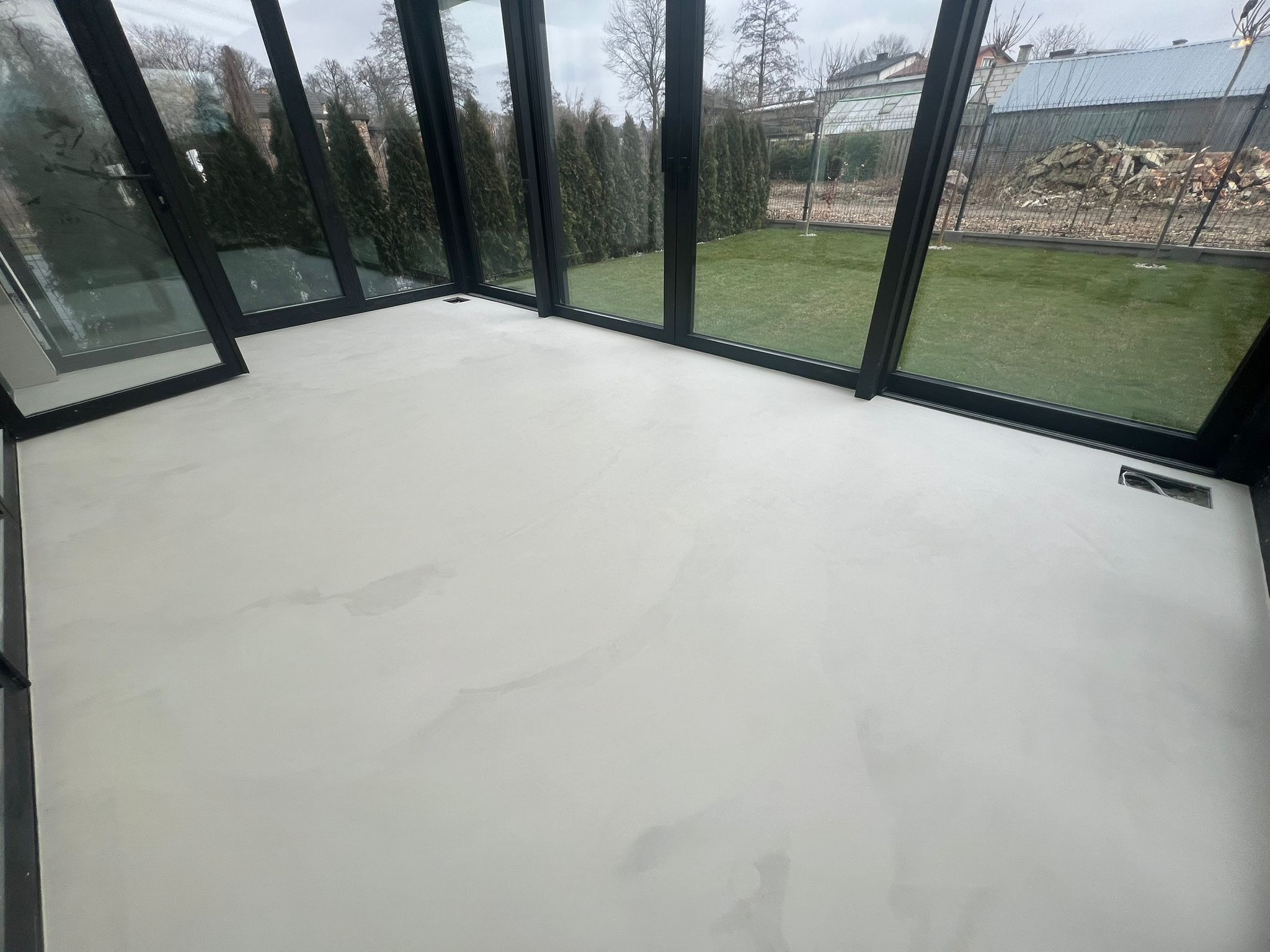 Jasna, gładka posadzka w oranżerii z czarnymi ramami okiennymi. Widok na zielony trawnik i drzewa za oknem. Minimalistyczny design, neutralna kolorystyka.