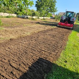 WYLĄG DREWNO BIOMASA ENERGIA - Niwelacja terenu: Traktor z glebogryzarką spulchnia ziemię na działce, przygotowując ją pod uprawę. Widoczne świeżo obrobione bruzdy i trawa w tle.