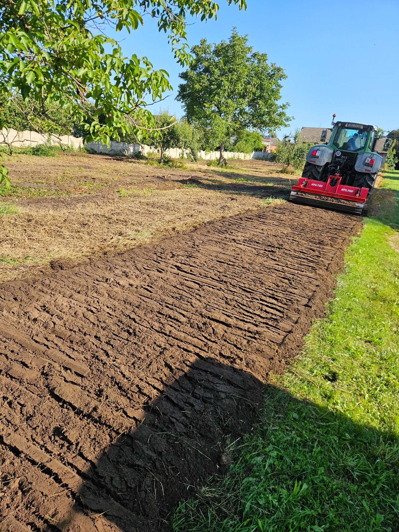 Niwelacja terenu: Traktor z glebogryzarką spulchnia ziemię na działce, przygotowując ją pod uprawę. Widoczne świeżo obrobione bruzdy i trawa w tle.