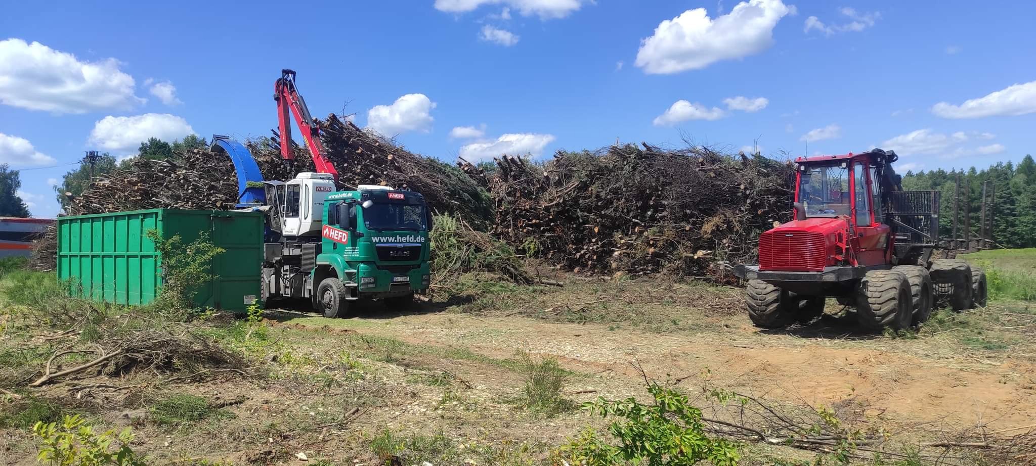 Ciężki sprzęt do wycinki drzew: zielony samochód z dźwigiem i czerwony traktor na tle sterty gałęzi i pni, błękitne niebo z chmurami w tle. Widok z poziomu gruntu.