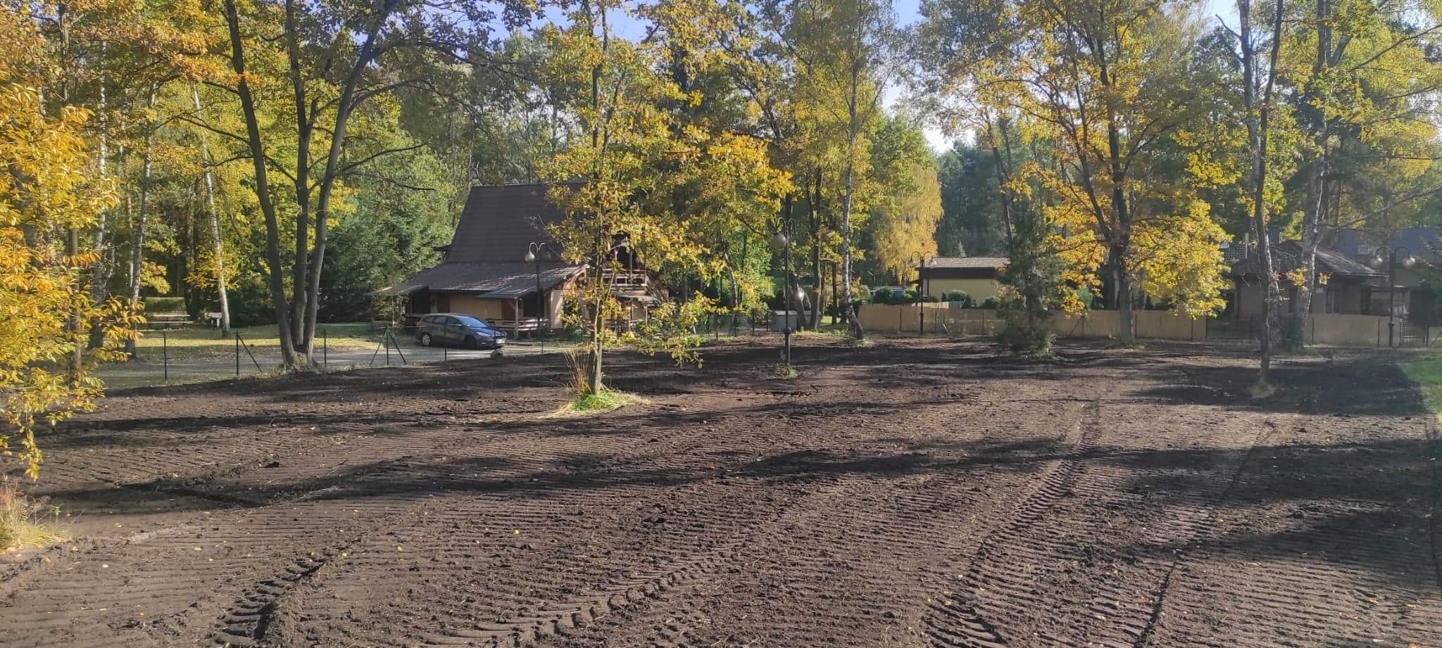 Przygotowany teren pod przyszły ogród, świeżo przekopana ziemia z widocznymi śladami opon, w tle dom z brązową dachówką i jesienne drzewa.