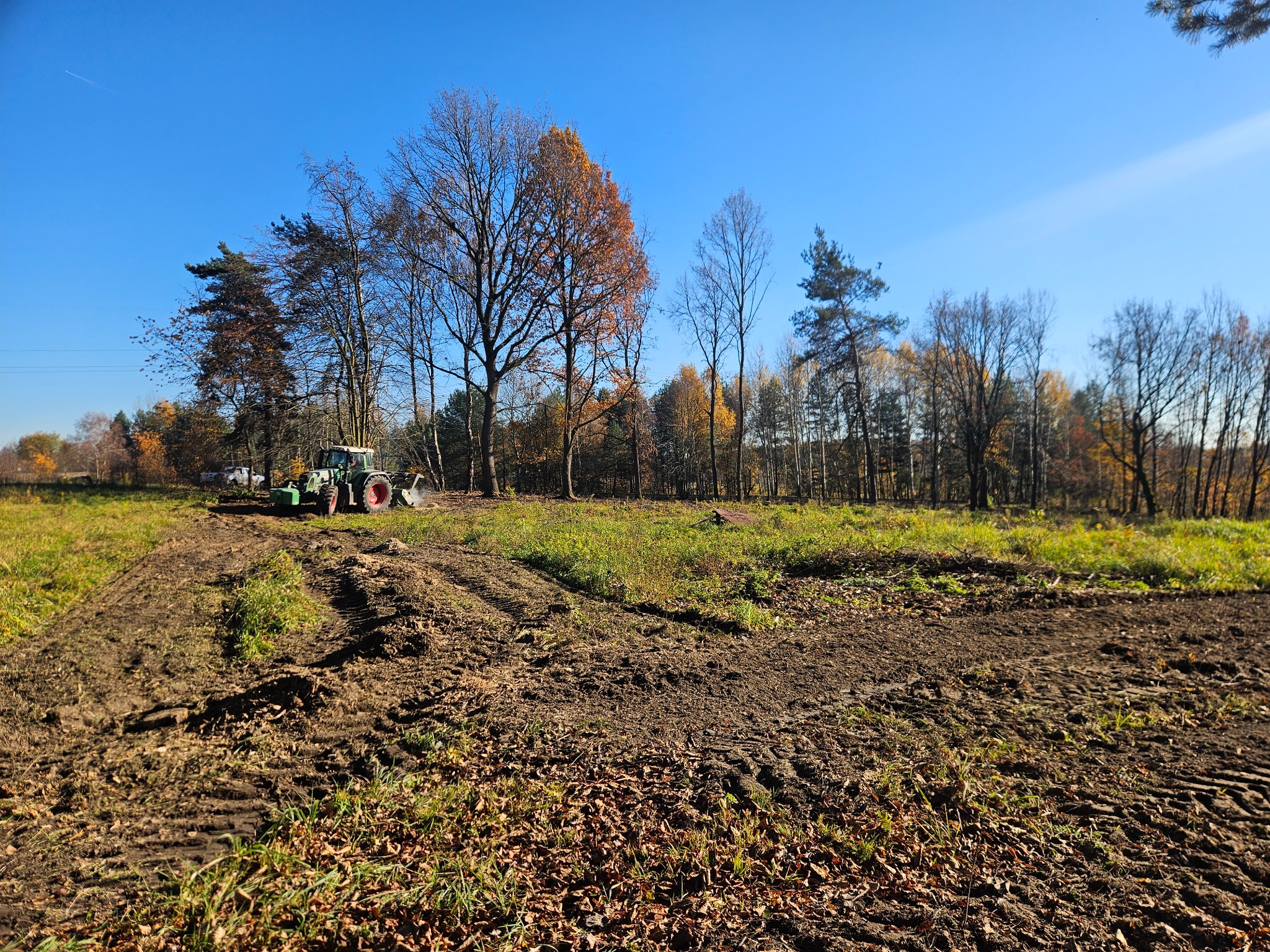 Traktor na polu po niwelacji terenu, widoczne ślady opon w ziemi, w tle drzewa z jesiennymi liśćmi i błękitne niebo. Słoneczny dzień.