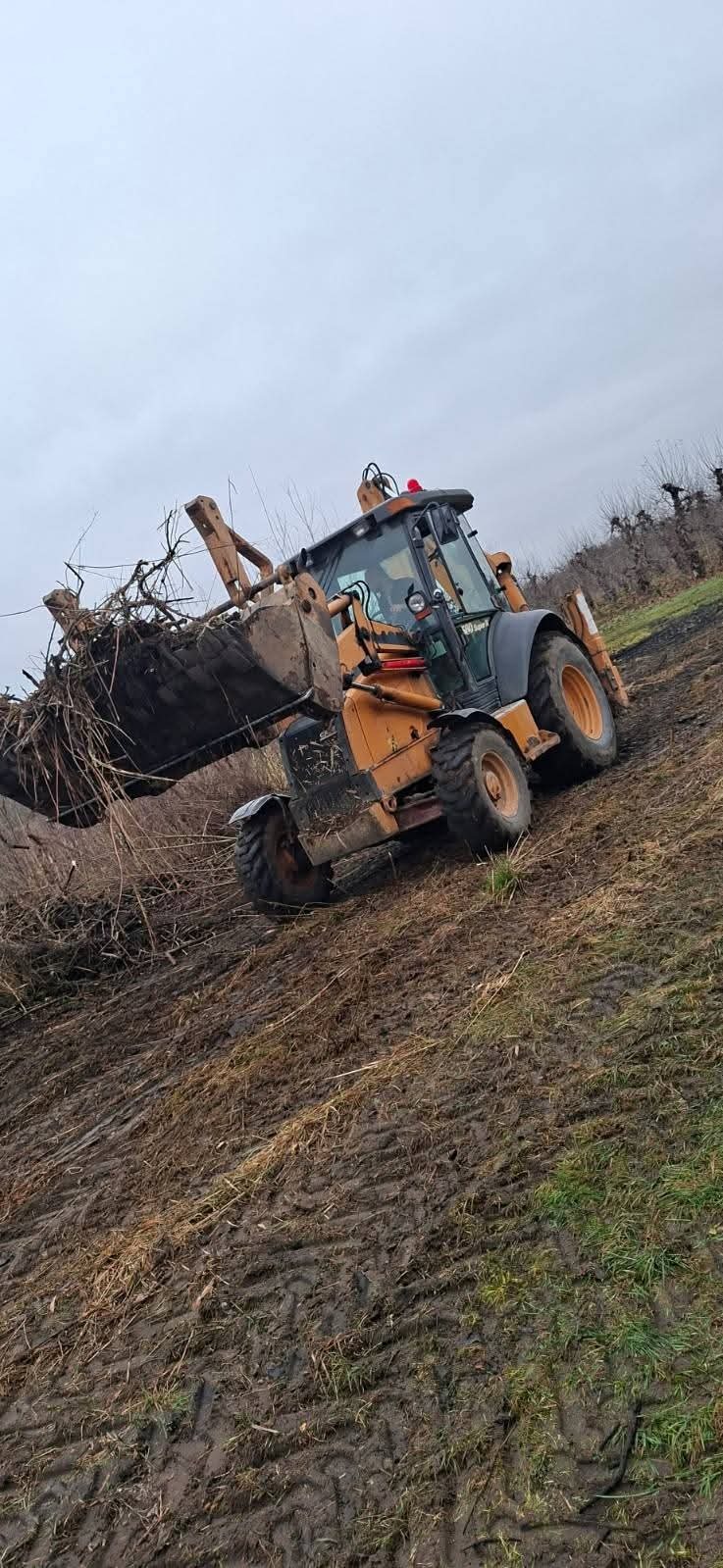 Żółto-szara koparko-ładowarka na błotnistym, pochylonym terenie, z pełną łyżką gałęzi i ziemi, pod pochmurnym niebem. Widoczne ślady opon w błocie.