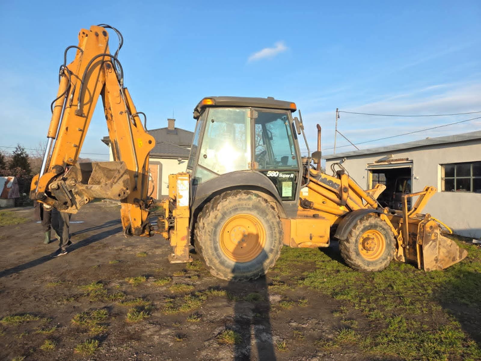 Żółta koparko-ładowarka Case 580 Super R na trawiastym terenie, z podniesionym ramieniem koparki i widocznym łyżką. W tle budynki i błękitne niebo.