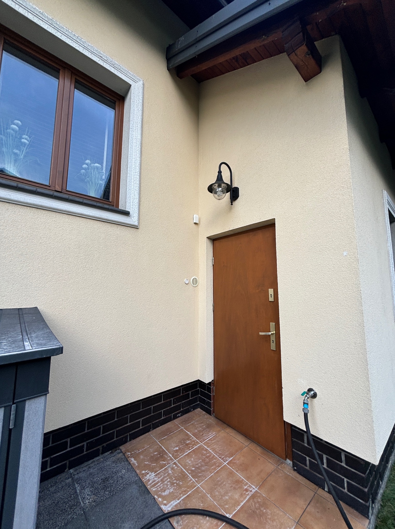 Elewacja domu w beżowym odcieniu z brązowymi oknami i drzwiami wejściowymi. Widoczny fragment kostki brukowej i węża ogrodowego. Oświetlenie zewnętrzne.