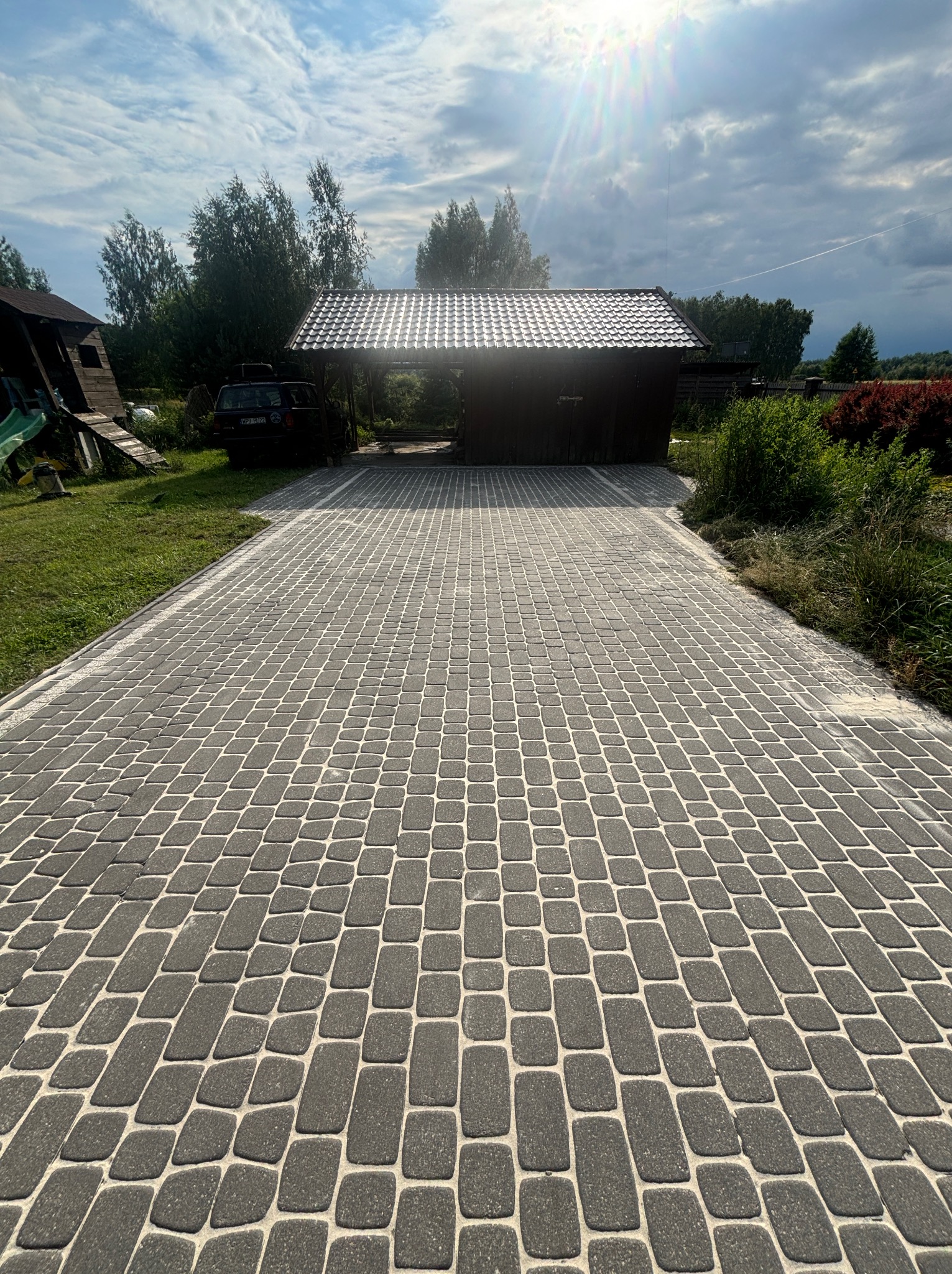 Ułożona, szara kostka brukowa na podjeździe, z widocznym budynkiem w tle i fragmentem samochodu. Słoneczny dzień, lekko pochmurne niebo.