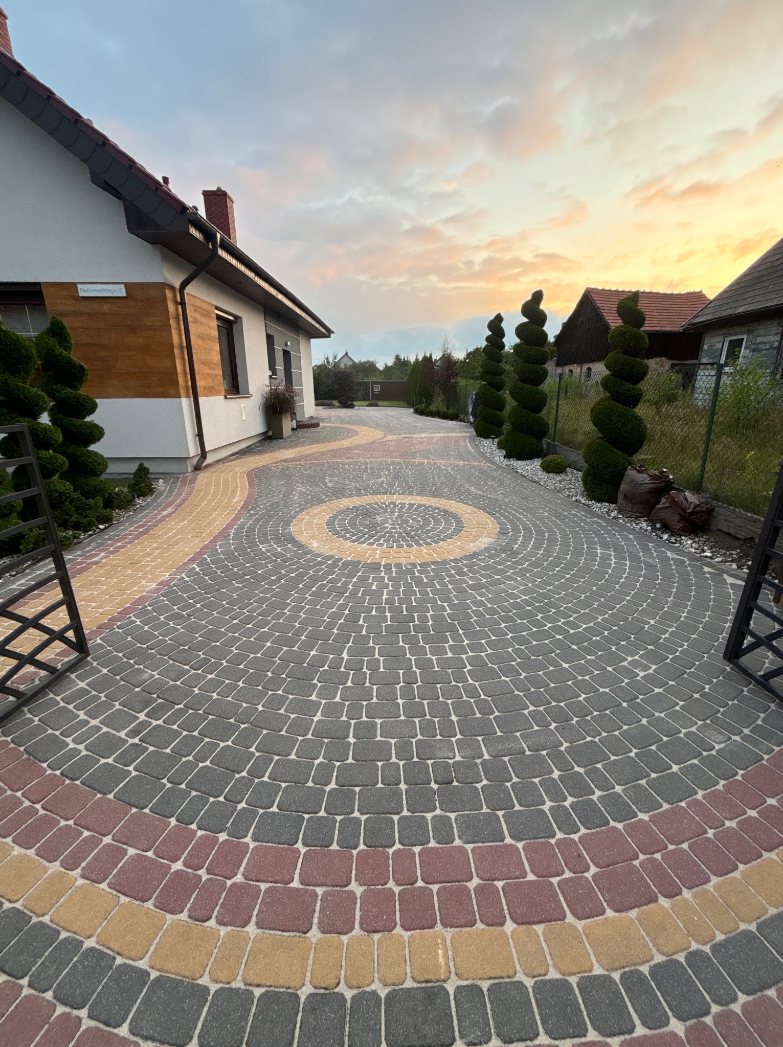 Kolorowa kostka brukowa ułożona w okrągły wzór na podjeździe domu jednorodzinnego, z ozdobnymi krzewami i wieczornym niebem w tle. Widok z poziomu gruntu.