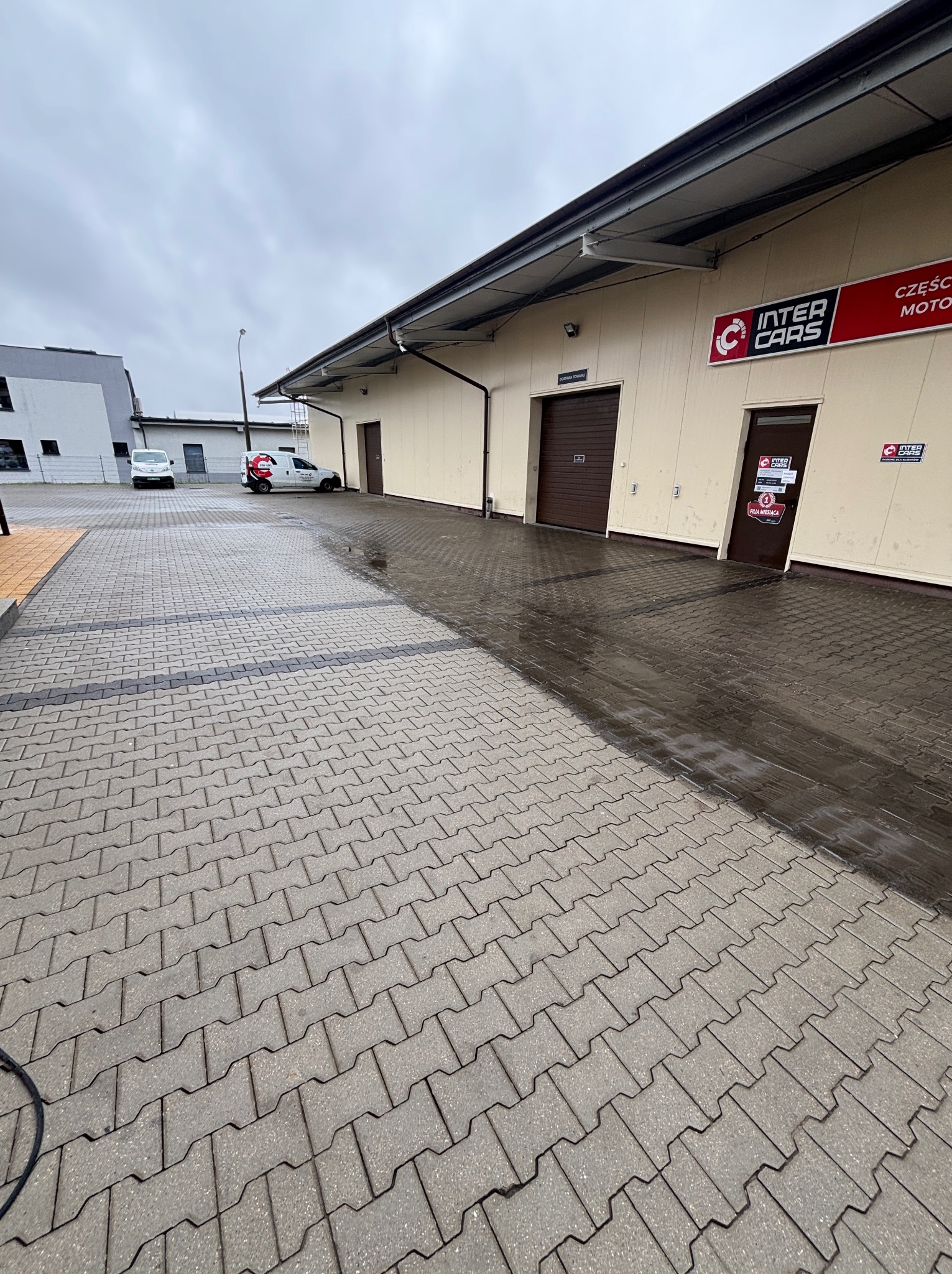 Częściowo umyta kostka brukowa przed budynkiem Inter Cars. Widoczna różnica w kolorze między czystą a brudną powierzchnią. Na drugim planie zaparkowane busy.