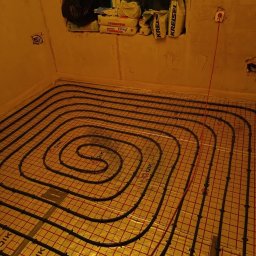 FILIP JĘDRZEJEWSKI - Instalacja ogrzewania podłogowego z czarnymi rurami ułożonymi spiralnie na złotej folii izolacyjnej QIK w surowym pomieszczeniu z workami budowlanymi na parapecie.