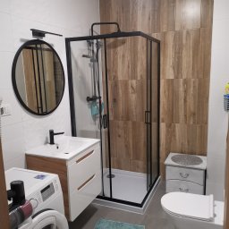 Glazurnik Jerzy Radtke - Nowoczesna łazienka po remoncie: kabina prysznicowa z czarną ramą, umywalka z szafką w stylu drewna, okrągłe lustro i płytki imitujące drewno na ścianie.