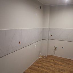 Glazurnik Jerzy Radtke - Fragment wnętrza w trakcie prac. Częściowo położone płytki imitujące marmur na ścianie, widoczne instalacje elektryczne i hydrauliczne, podłoga wyłożona panelami.