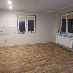 Glazurnik Jerzy Radtke - Jasne panele podłogowe imitujące drewno w nowym wnętrzu. Częściowo wykończona ściana z białymi płytkami. Dwa okna z roletami. Świeżo pomalowane ściany na szaro.