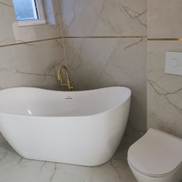 Glazurnik Jerzy Radtke - Biała, wolnostojąca wanna i toaleta w nowoczesnej łazience z marmurowymi płytkami i złotymi akcentami. Minimalistyczny design, jasne wnętrze.