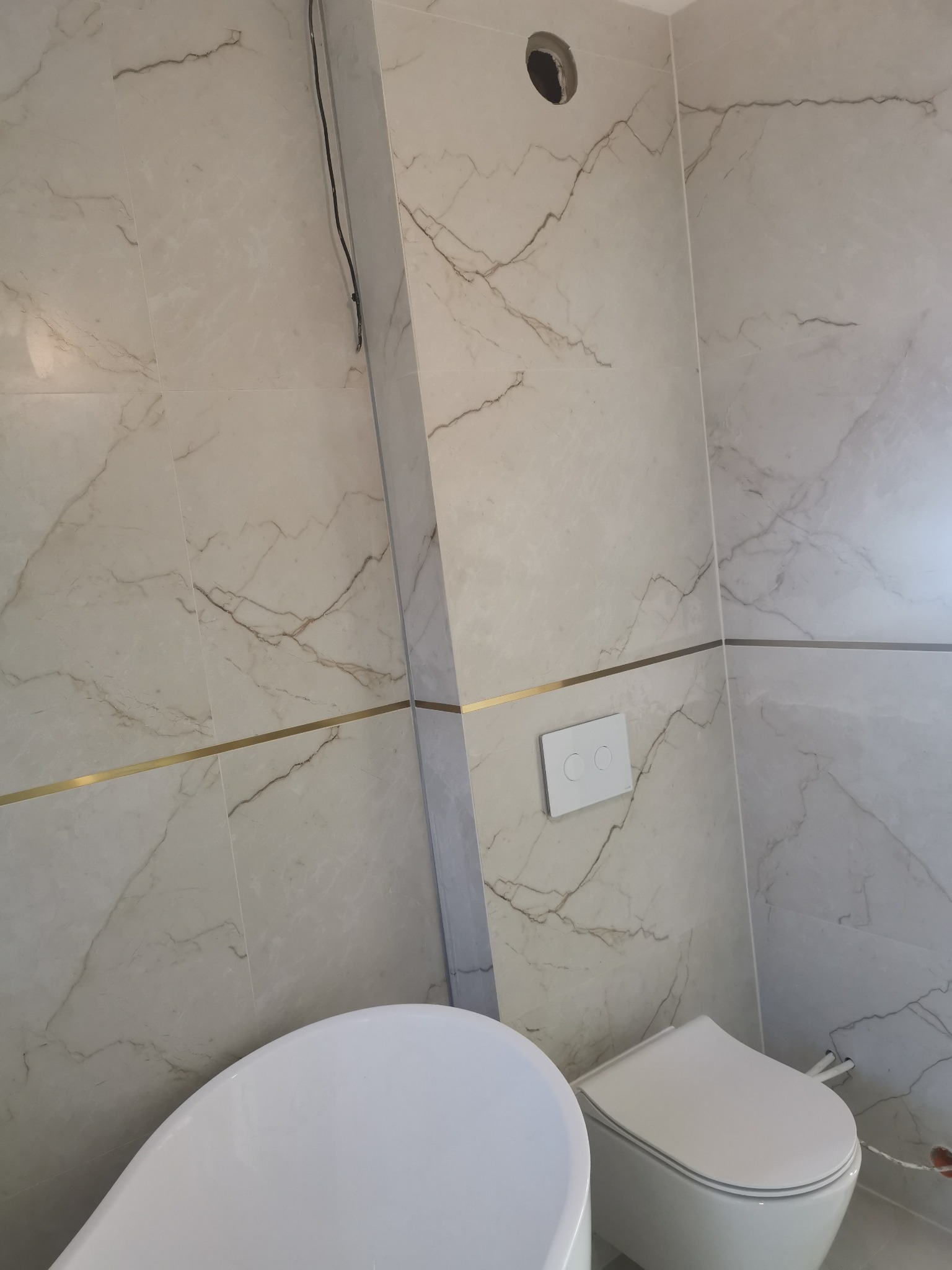 Elegancka łazienka z marmurowymi płytkami, złotymi akcentami i nowoczesną armaturą. Widoczna wanna, toaleta i przycisk spłuczki. Otwór w ścianie.