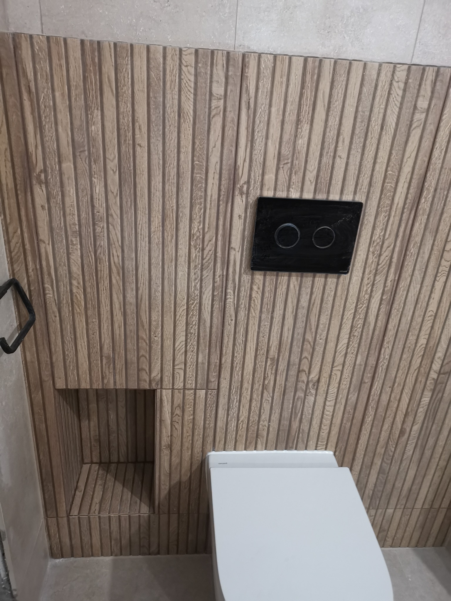 Nowoczesna toaleta podwieszana z drewnopodobną okładziną ścienną z ryflowanym wzorem i czarnym przyciskiem spłukującym. W ścianie wnęka wykończona tym samym materiałem.