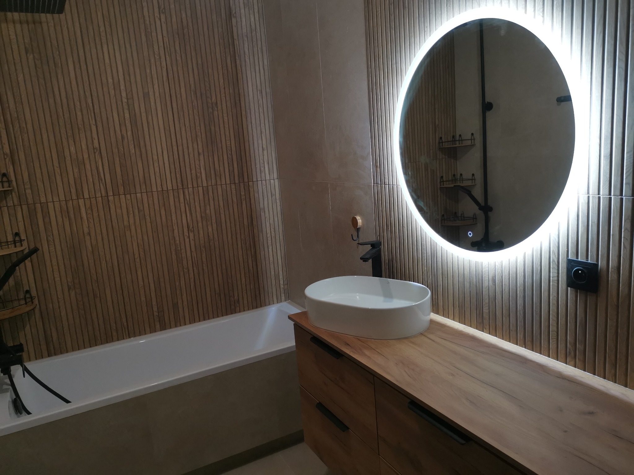 Nowoczesna łazienka z drewnianą okładziną ścienną, owalnym lustrem z podświetleniem LED i białą umywalką nablatową na drewnianej szafce. Minimalistyczny design.