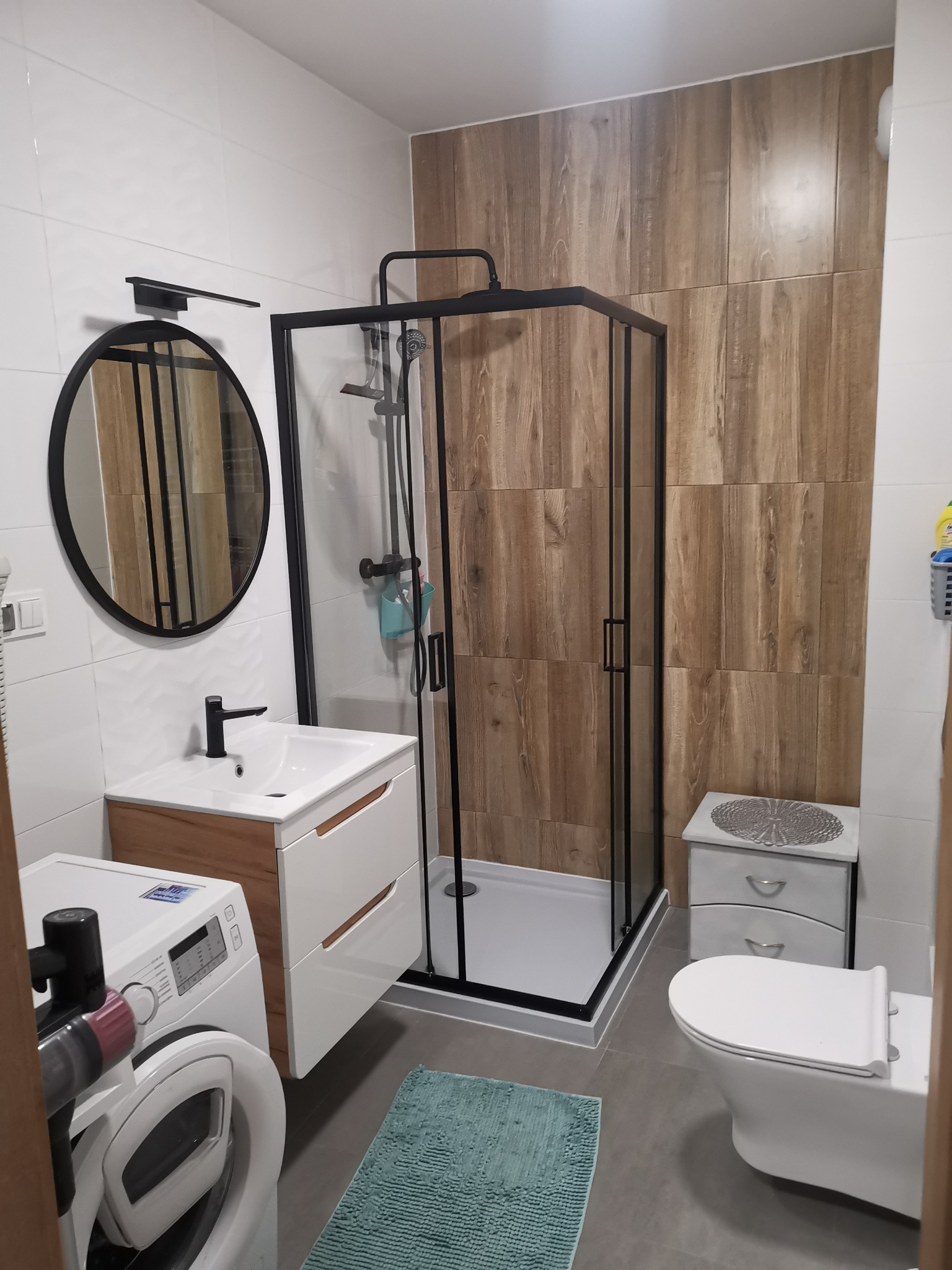 Nowoczesna łazienka po remoncie: kabina prysznicowa z czarną ramą, umywalka z szafką w stylu drewna, okrągłe lustro i płytki imitujące drewno na ścianie.