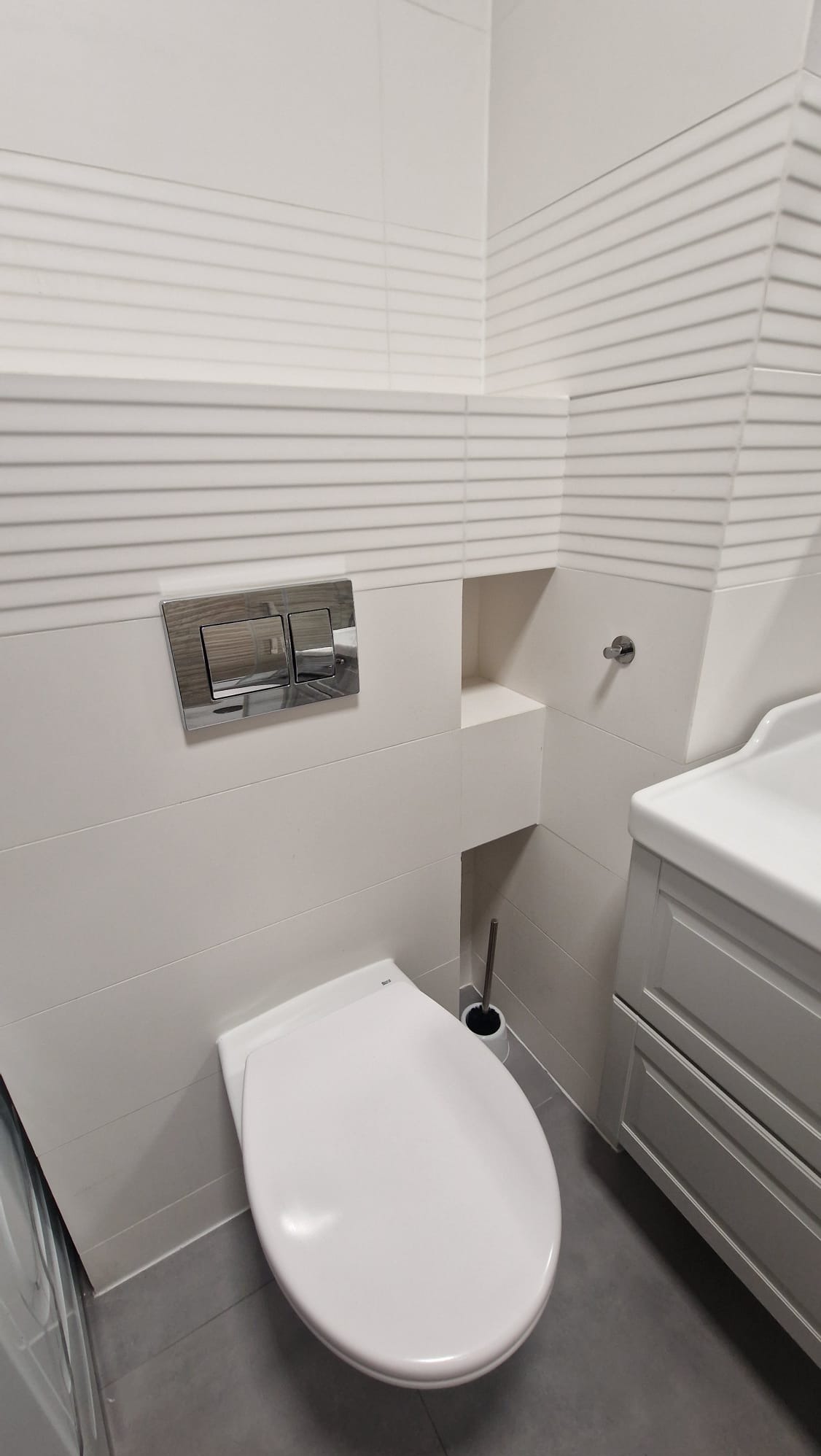 Nowoczesna toaleta podwieszana z chromowanym przyciskiem spłukującym i dekoracyjnymi płytkami w paski. W tle szafka umywalkowa i wnęka w ścianie.