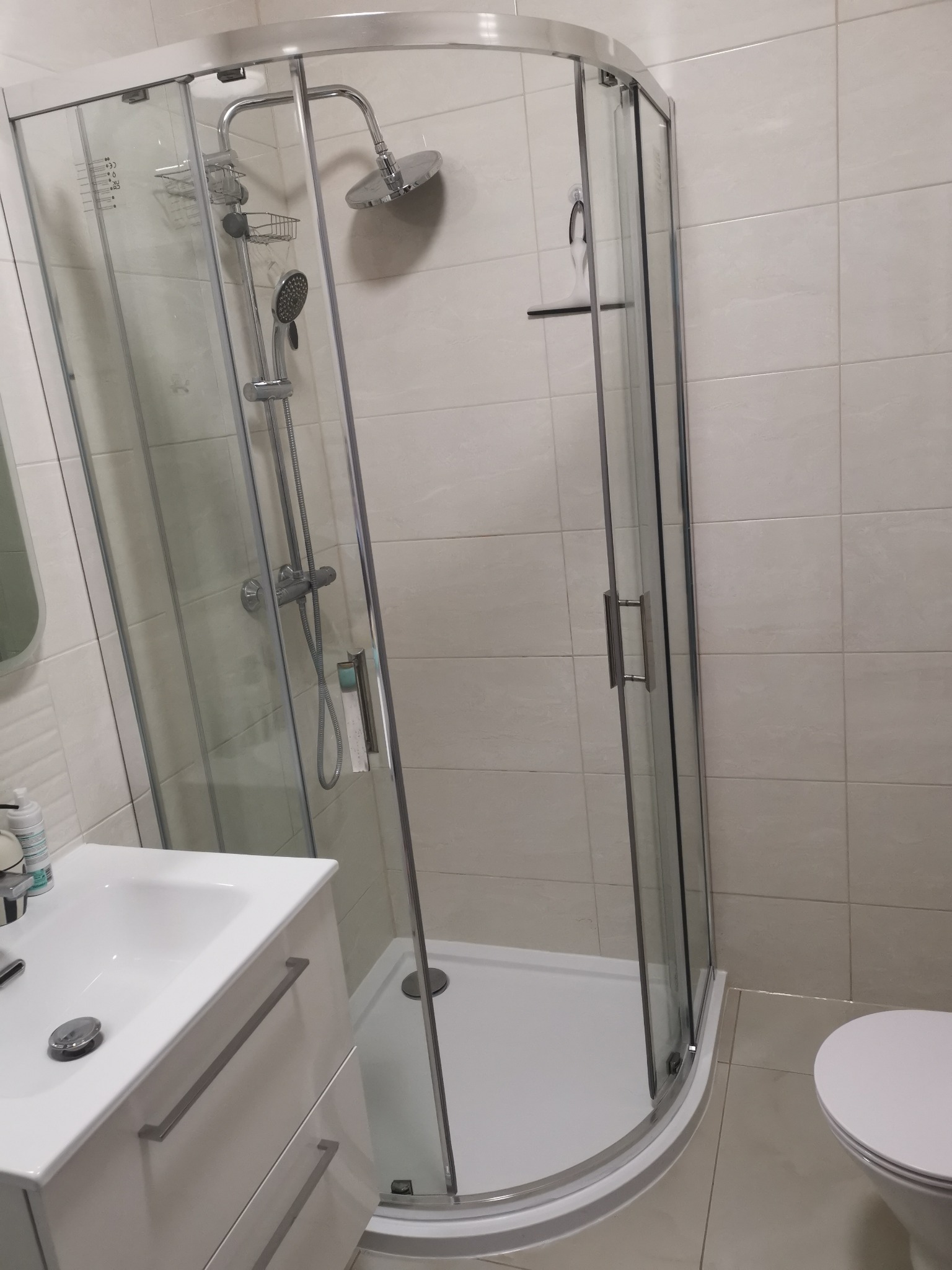 Nowoczesna kabina prysznicowa narożna z przesuwnymi drzwiami, zamontowana w łazience wyłożonej jasnymi płytkami. Widoczna umywalka z szafką i fragment toalety.